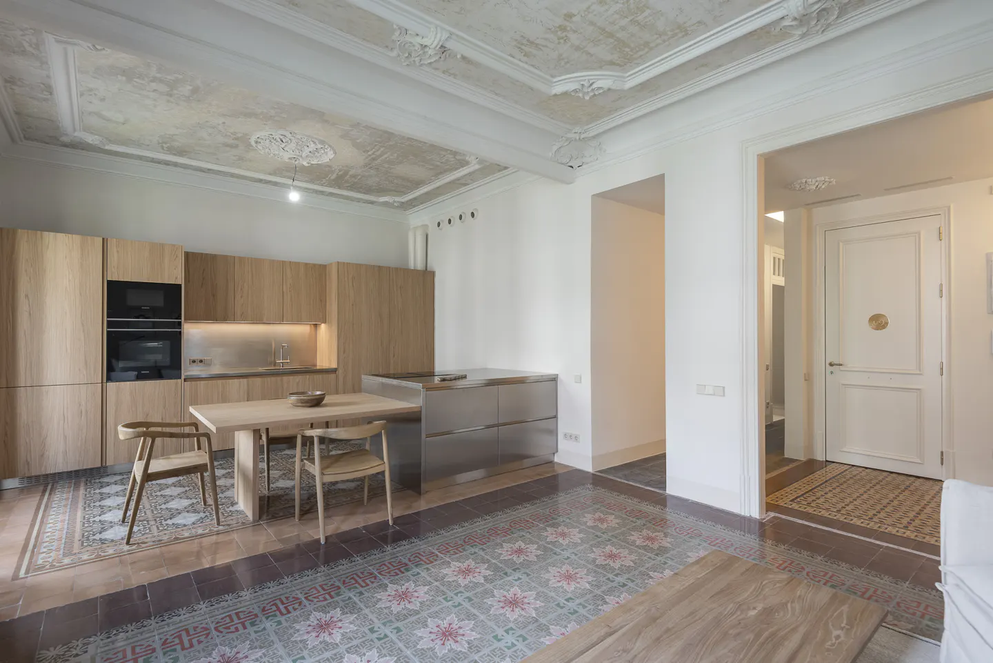 A bright kitchen with light wood cabinets, stainless steel island, and a table with two chairs. The floor has patterned tiles and the ceiling has decorative molding.