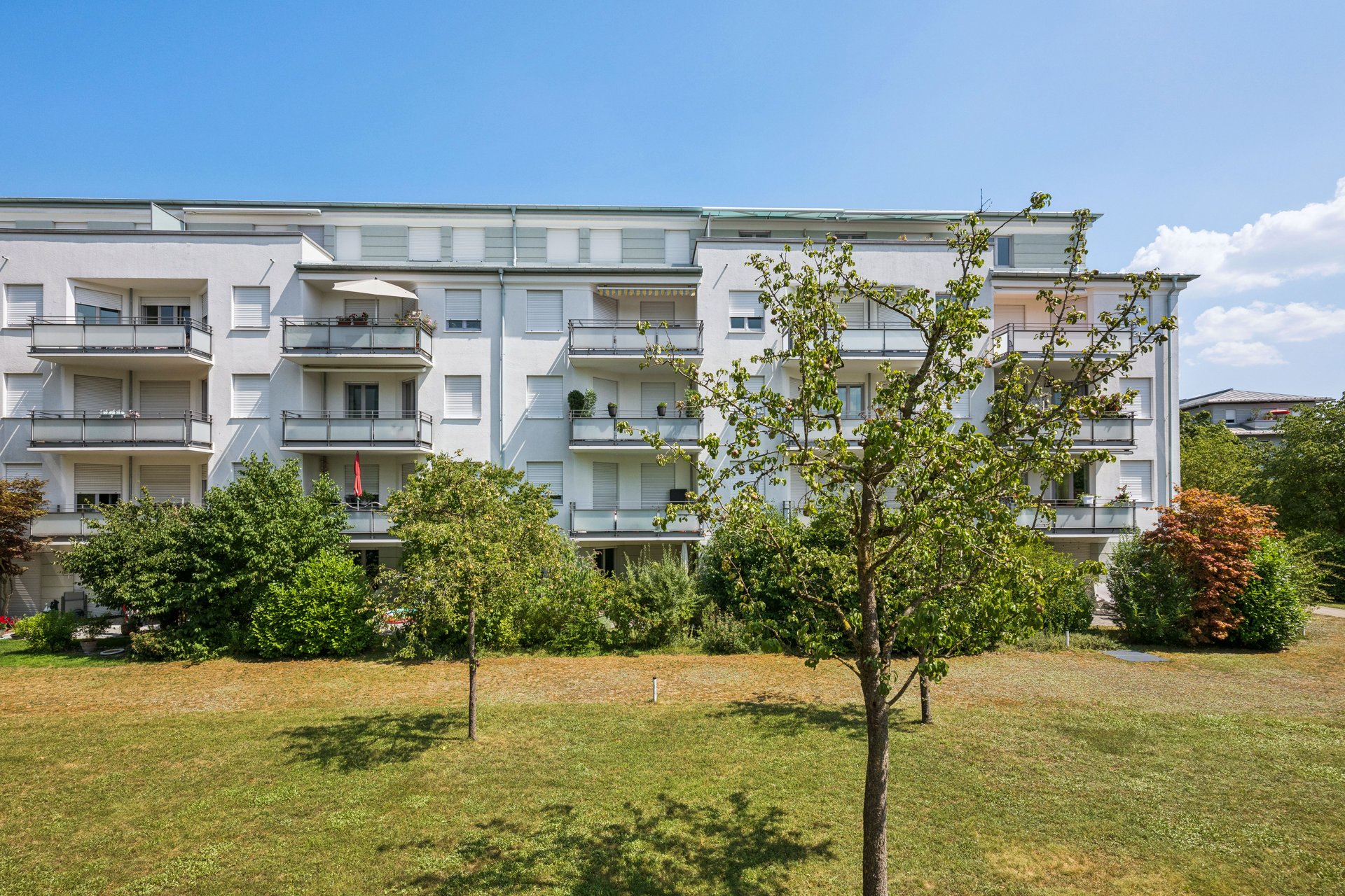 Modern white apartment building with balconies and green trees in front, set against a clear blue sky. [AI-generated text]