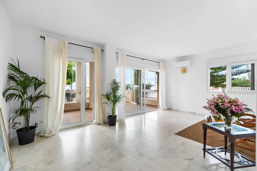 Bright, airy living room with marble floors, white walls, and balcony access. Plants, flowers, and a glass table add warmth.