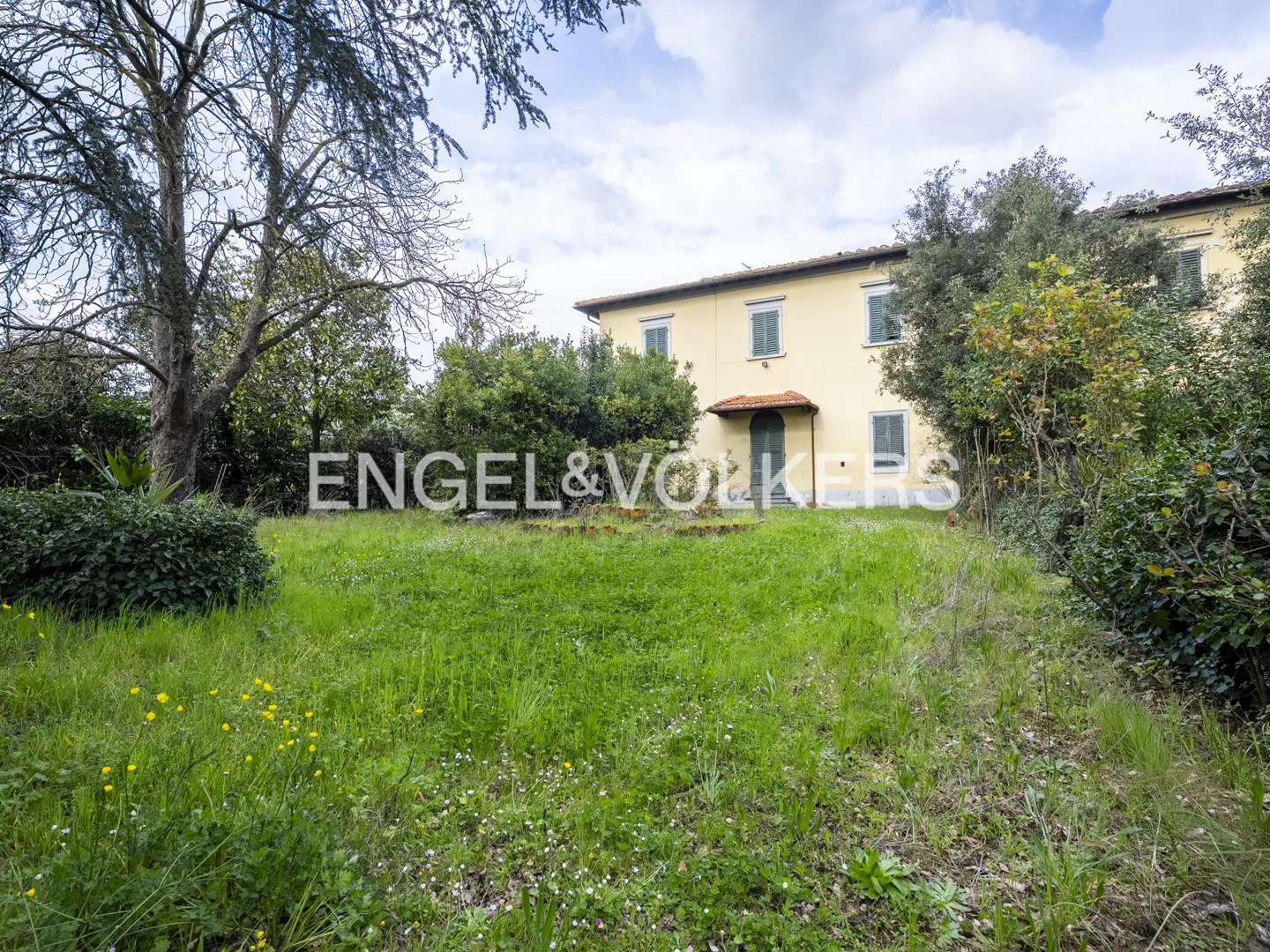 Exterior view of a two-story yellow house with green shutters and a red-tiled porch roof, surrounded by a green lawn and trees.