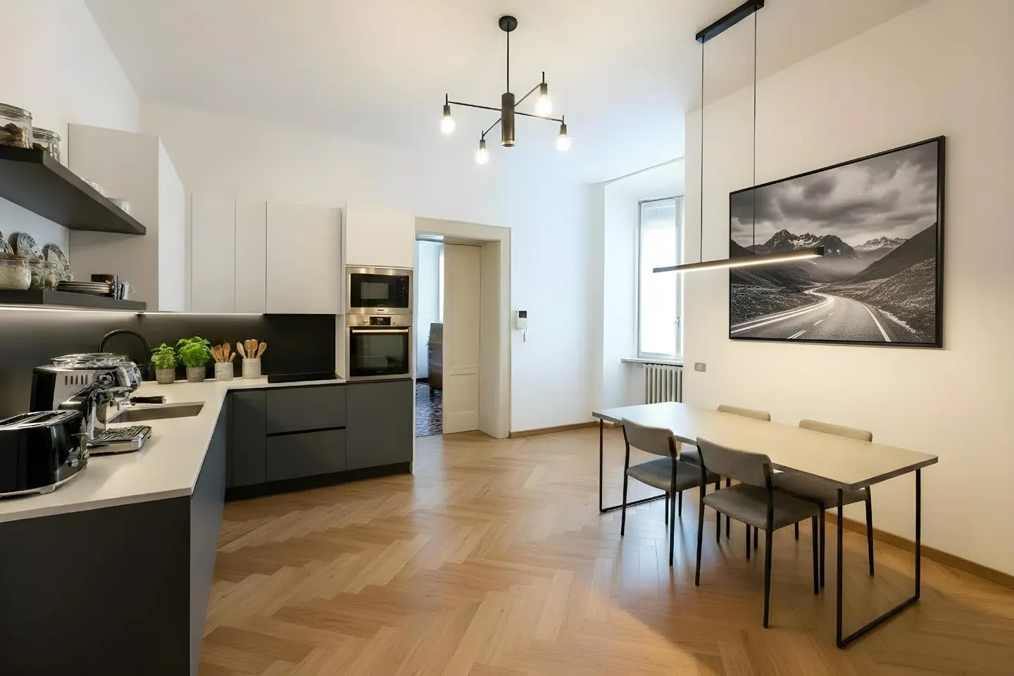 A modern kitchen with gray cabinets, white countertops, and a dining table with four chairs. A black and white mountain landscape art hangs on the wall.