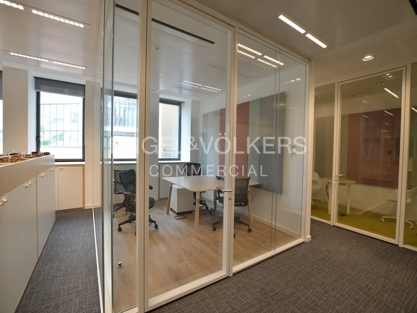 Office space with glass-walled rooms, gray carpet, and bright lighting. A table and chairs are visible inside one room.