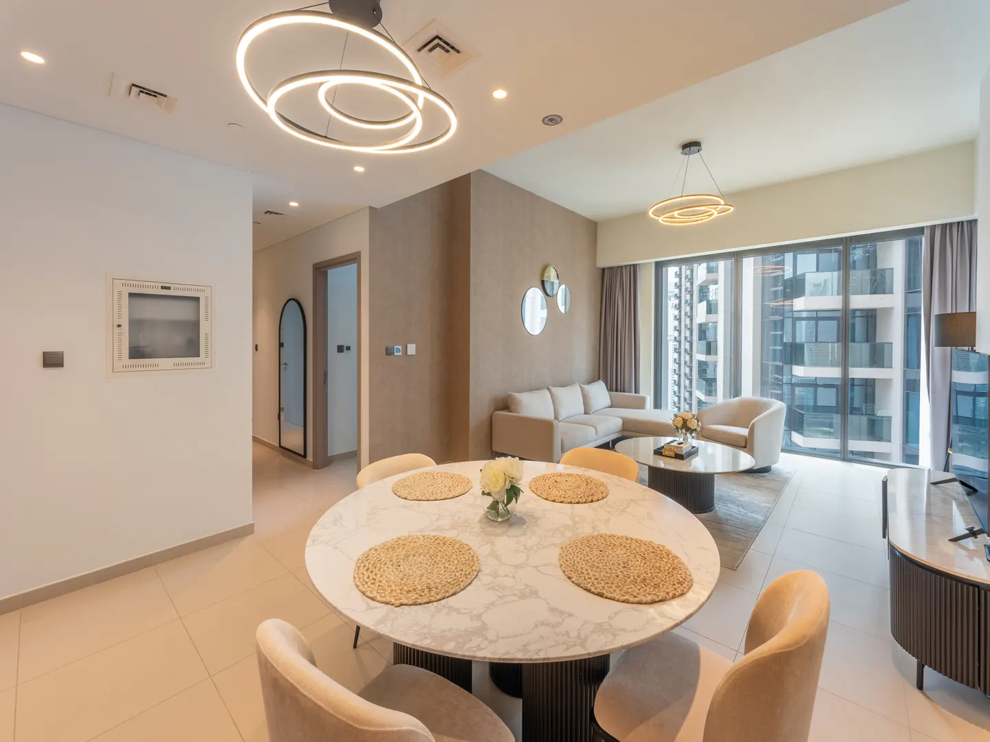 A modern apartment interior with a round marble table, beige chairs, and a living room with a large window.