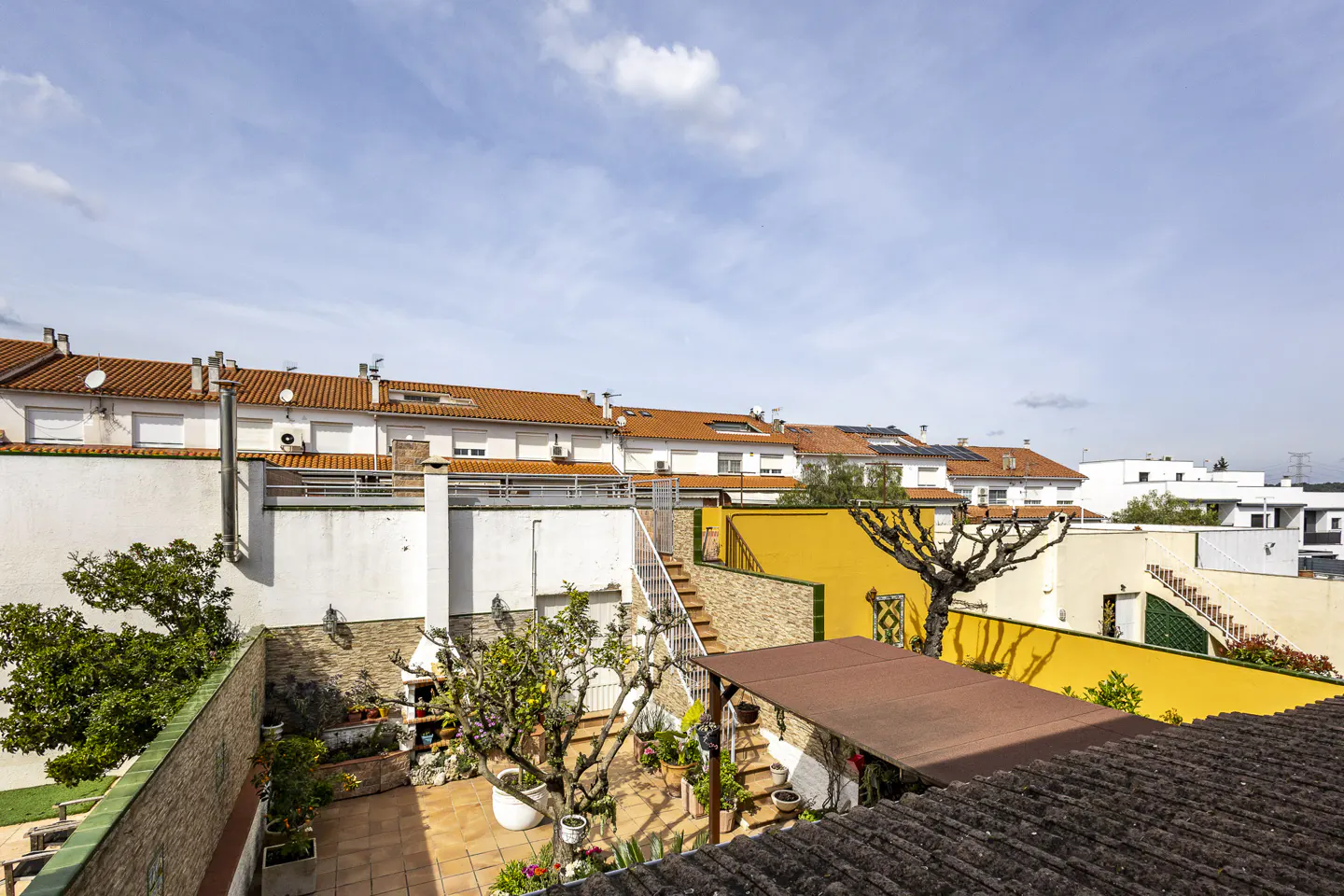 A high-angle view of a backyard with a tiled patio, plants, and a yellow wall, under a blue sky. Row houses with orange roofs are in the background.