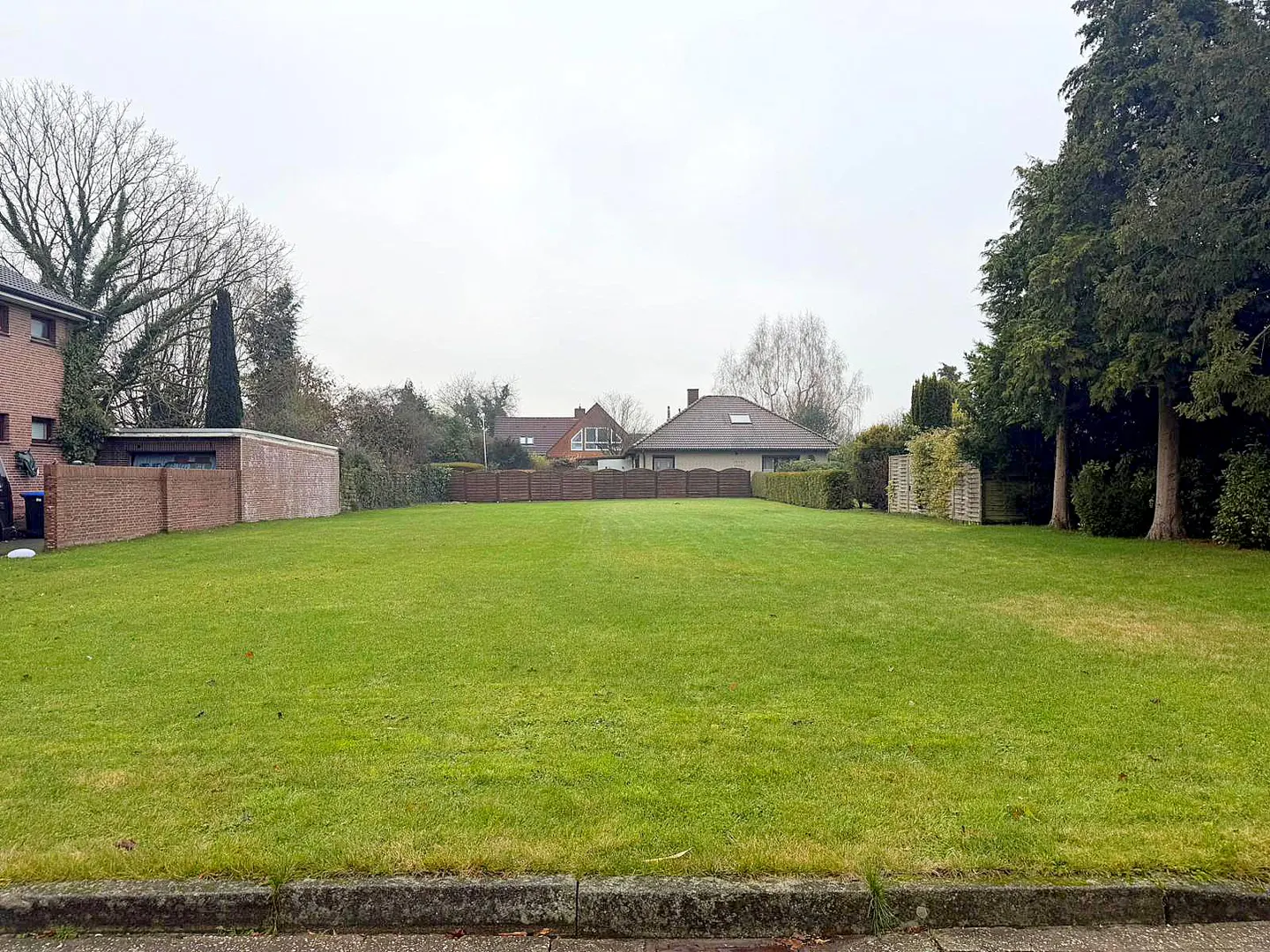 A vacant, rectangular lot with green grass, surrounded by brick walls, hedges, and trees under a cloudy sky.