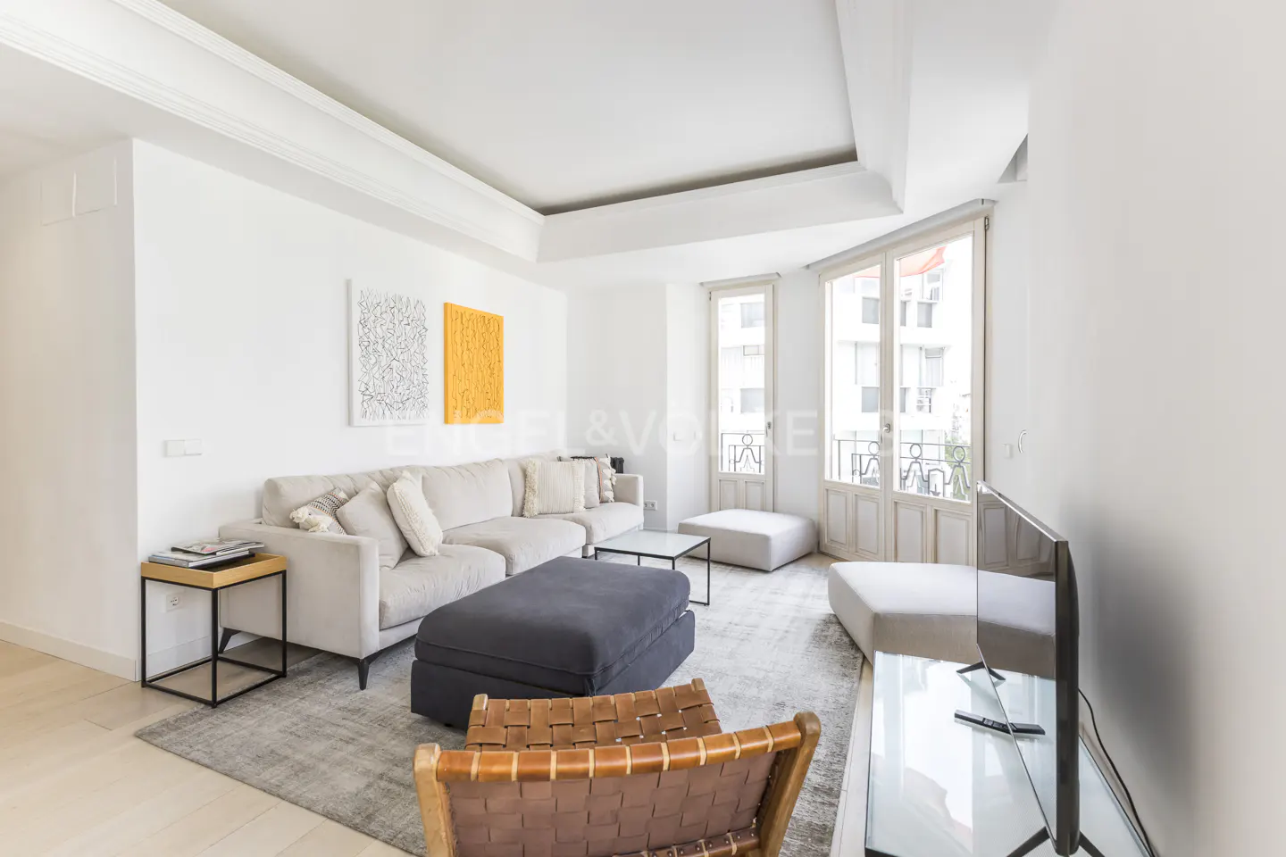 Bright living room with white walls, a beige sectional sofa, a gray rug, and a brown leather chair. Two colorful paintings hang on the wall.