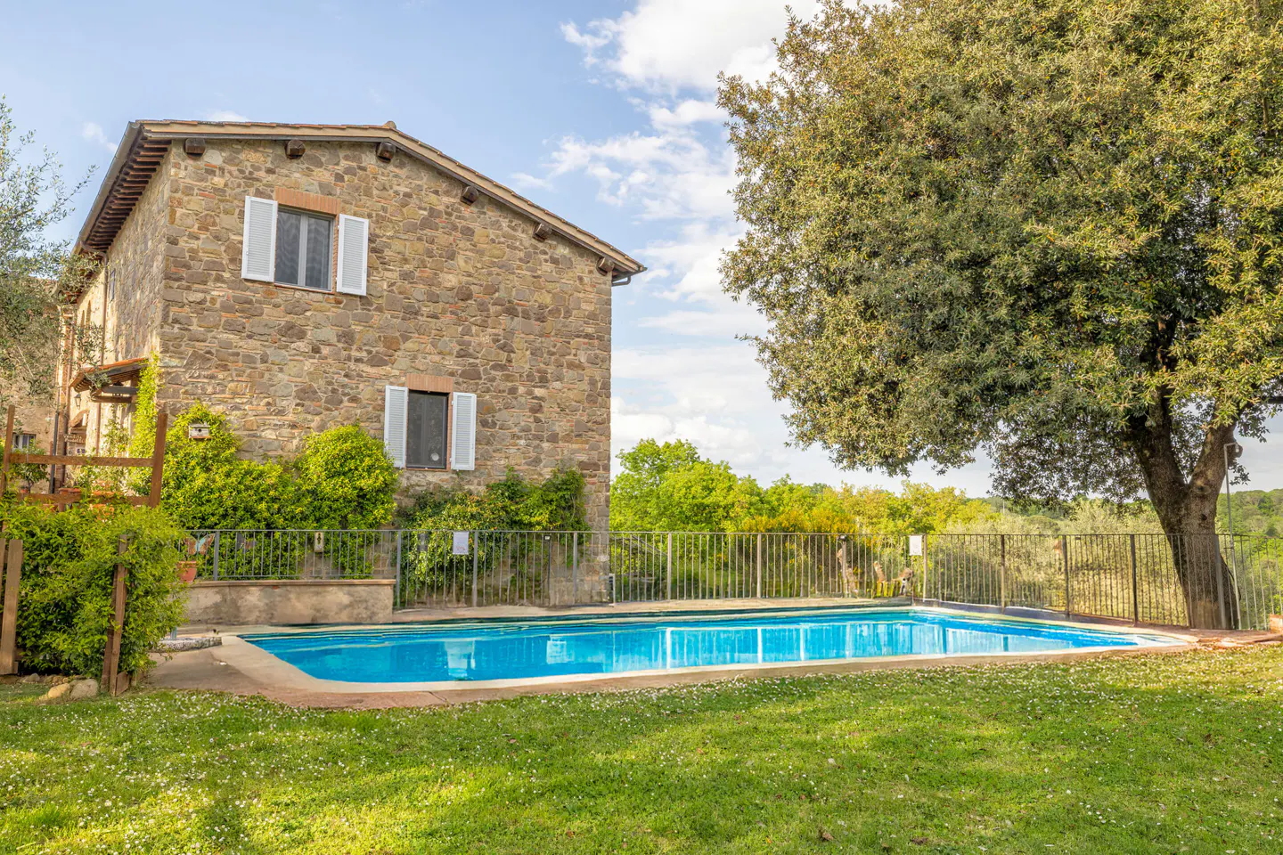 Stone house with white shuttered windows overlooks a blue tiled pool surrounded by green lawn and trees.