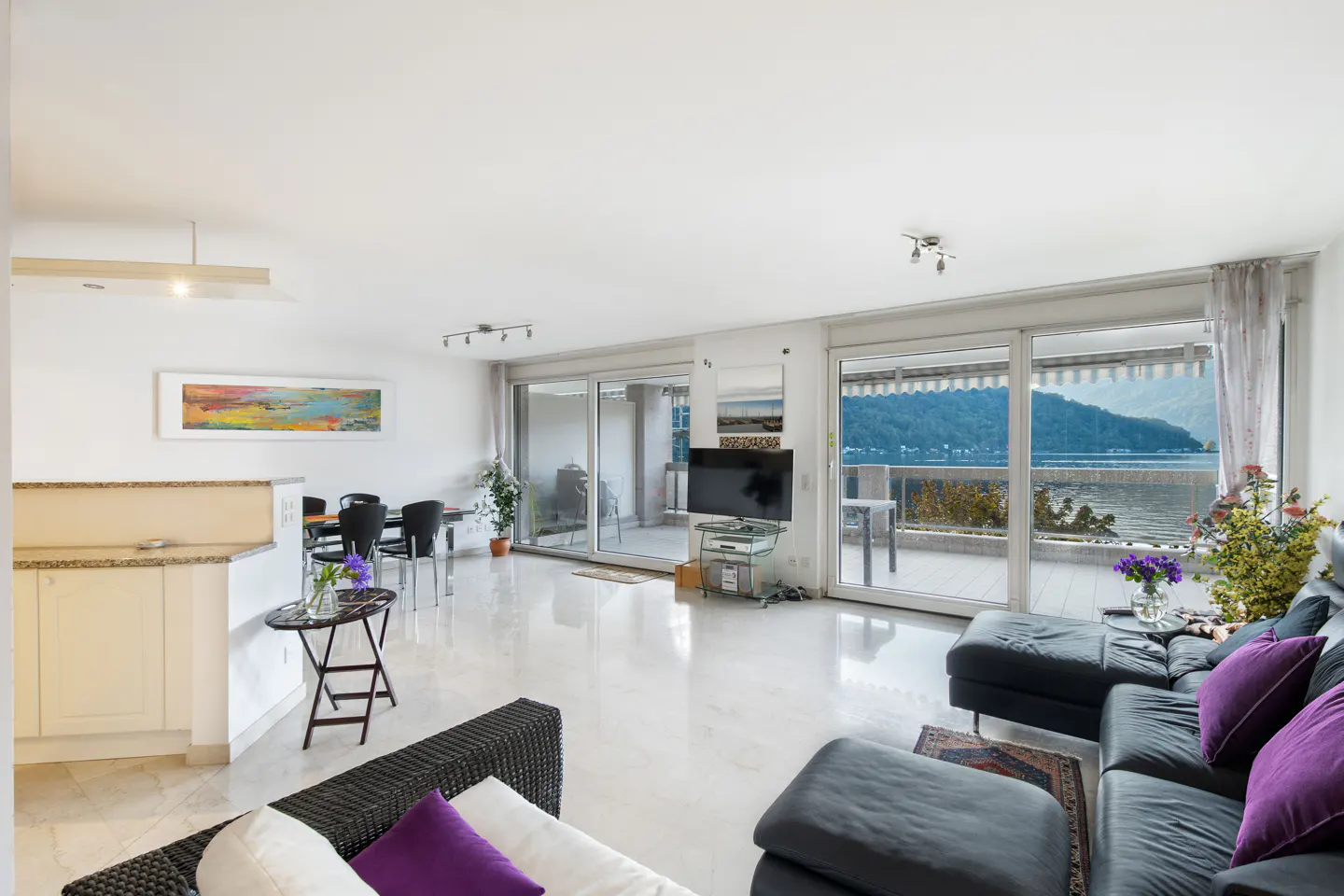Bright, open living room with marble floors, black leather sofa with purple pillows, and a view of a lake through sliding glass doors.