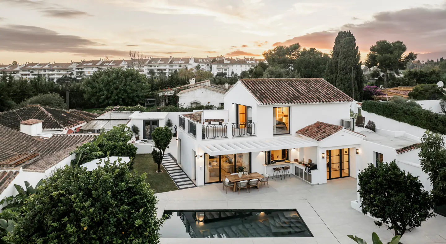 Aerial view of a white villa with a terracotta roof, swimming pool, outdoor dining area, and balcony at sunset.