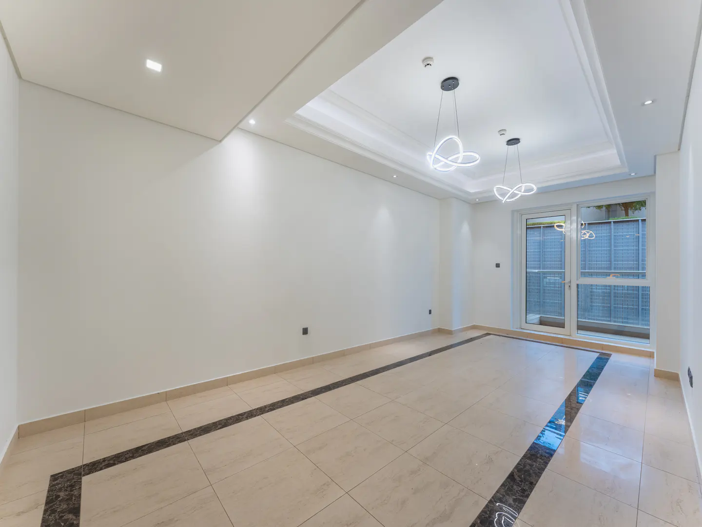 Bright, empty room with white walls, beige tile floor with dark border, and modern light fixtures. Glass doors lead to an outdoor area.