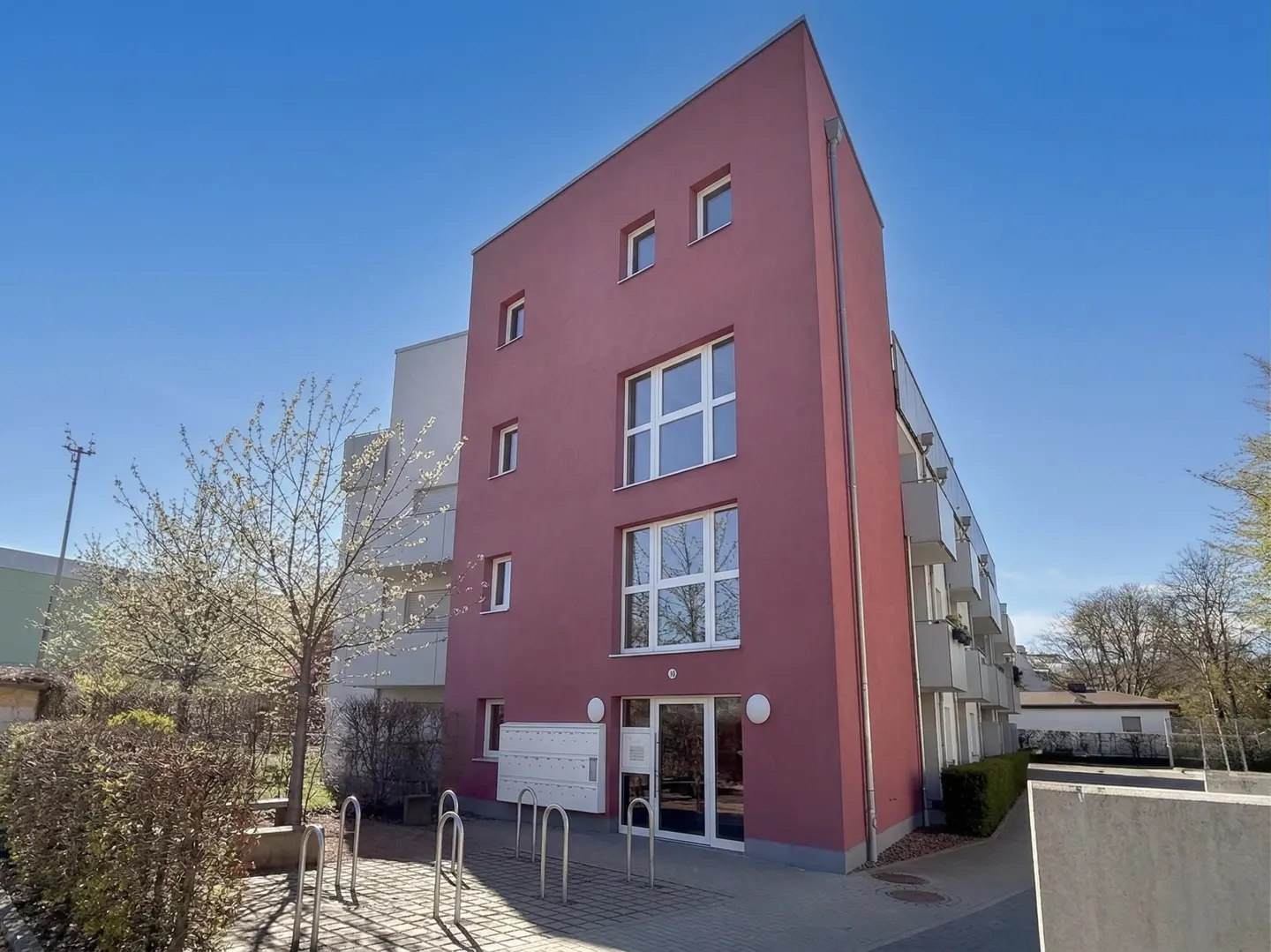 Exterior view of a modern, multi-story apartment building with a red facade and white-framed windows under a clear blue sky.