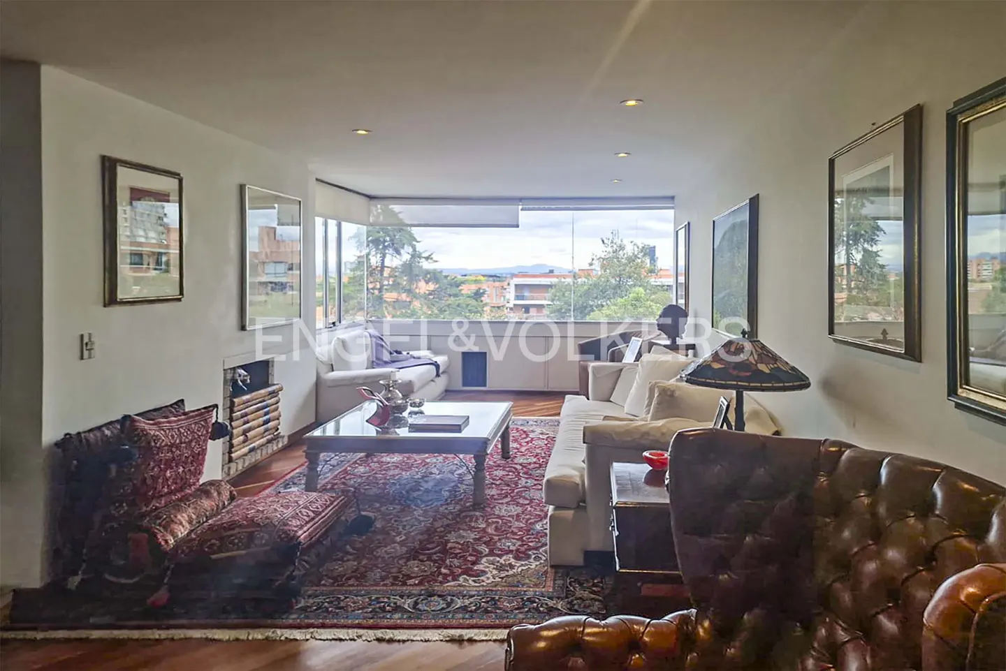 A living room with white walls, hardwood floors, and a red patterned rug. There are two sofas, a coffee table, and a large window with a view of trees and buildings.
