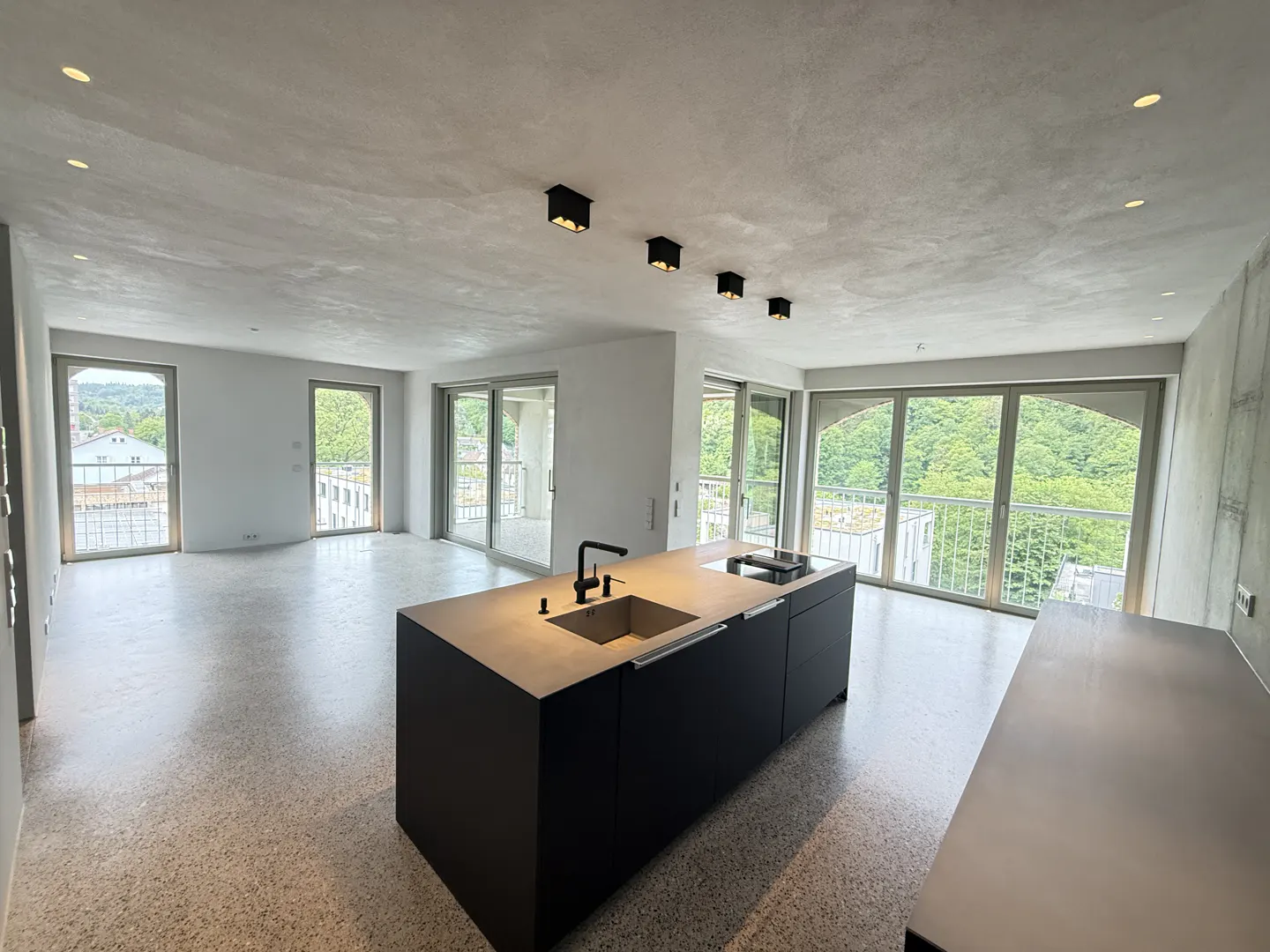 A modern apartment interior with a black kitchen island, large windows, and a view of greenery.