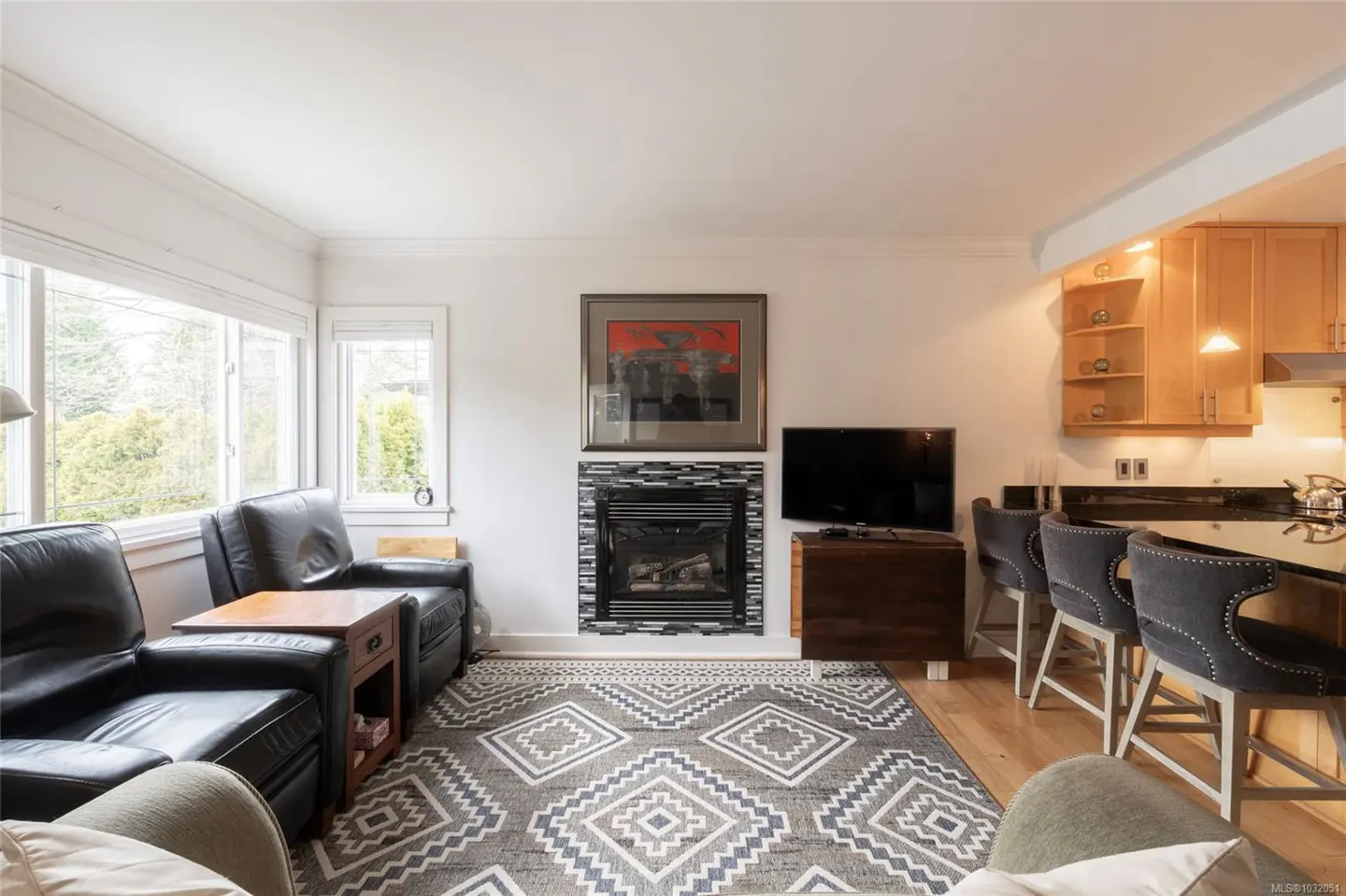 Living room with black leather chairs, fireplace, TV, and kitchen bar with gray stools. A gray patterned rug is on the wood floor.