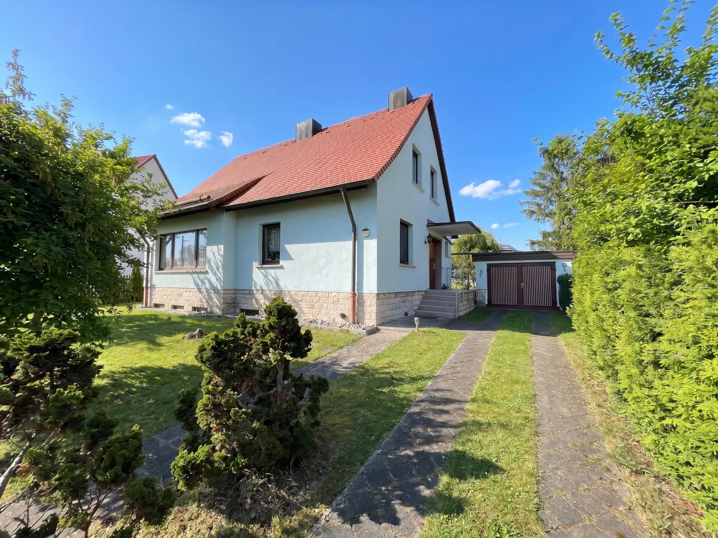 Exterior view of a light blue house with a red roof, green lawn, and a stone driveway leading to a garage.