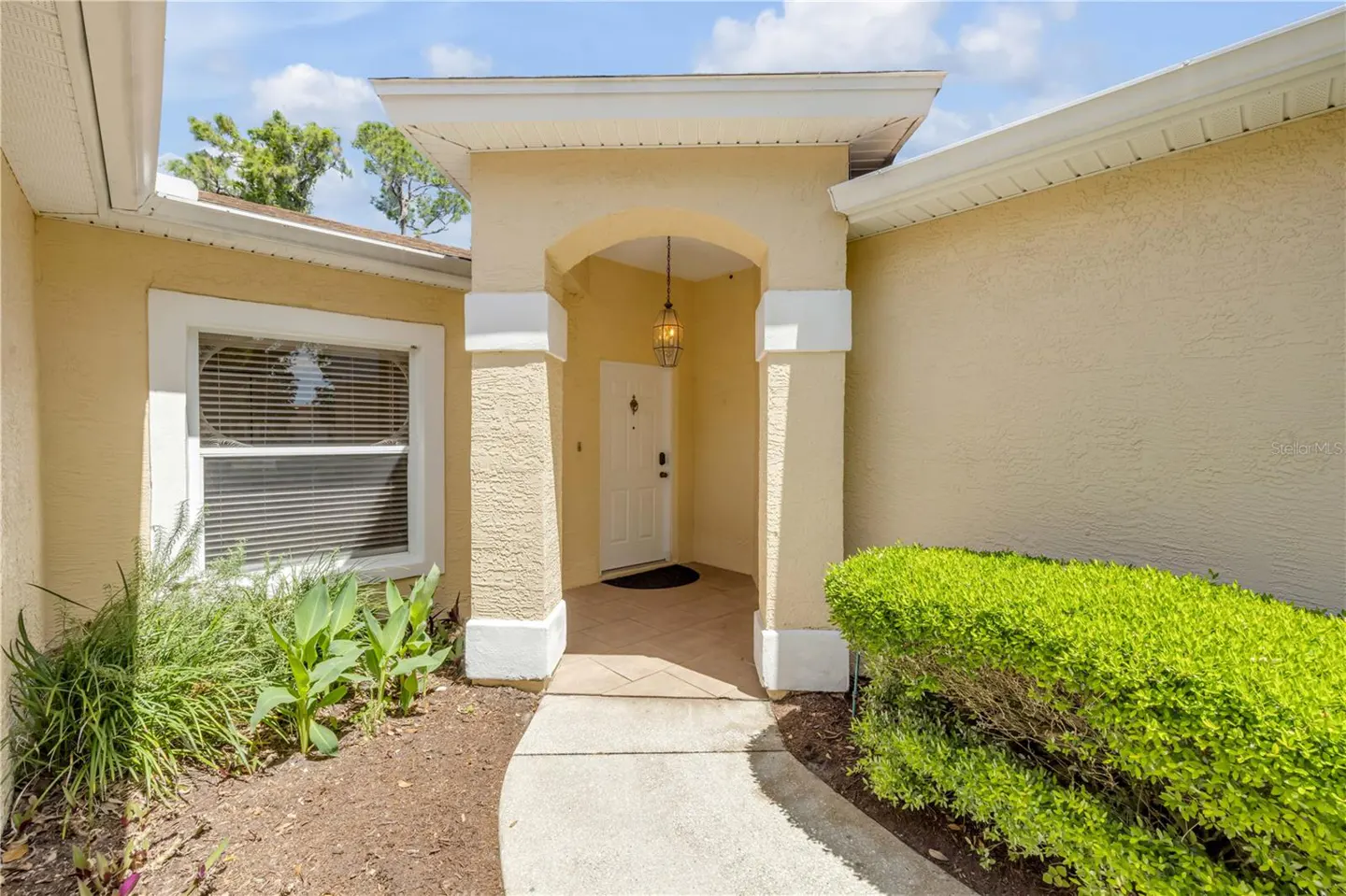 Exterior view of a beige stucco house with a white front door under an arched porch with white columns. A concrete walkway leads to the entrance.