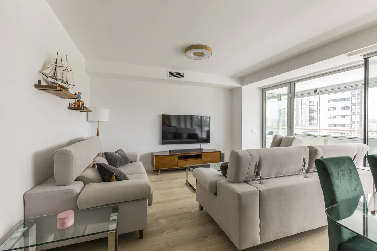 Bright living room with two beige sofas, a wooden TV stand with a black TV, and a glass coffee table. Large windows overlook a city view.