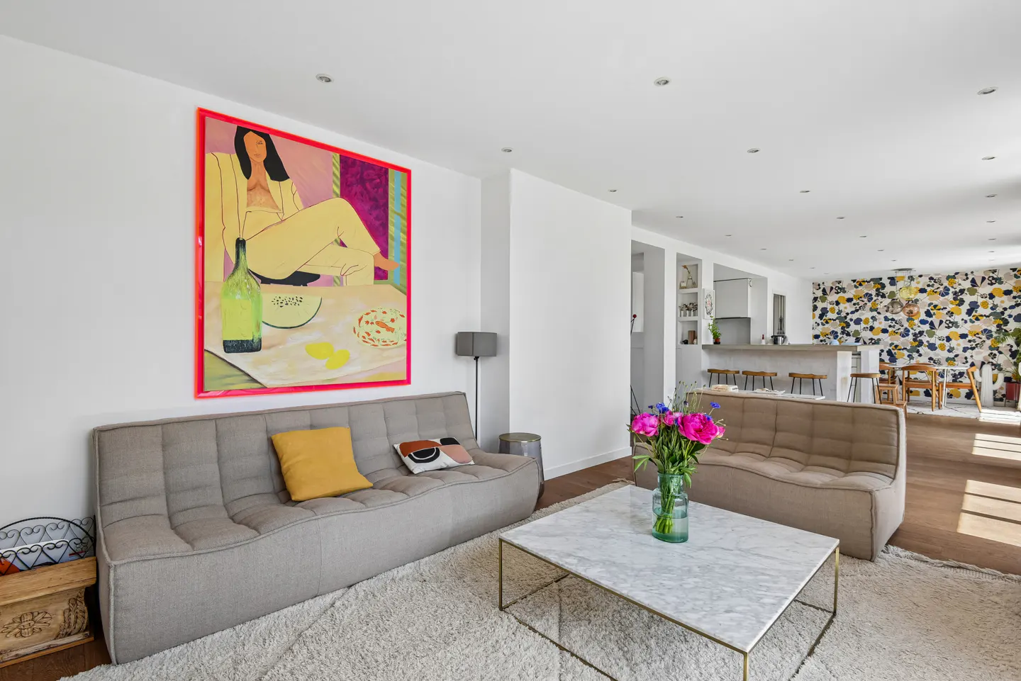 Modern living room with beige sofas, marble coffee table, bright floral art, and an open kitchen with a patterned wall.