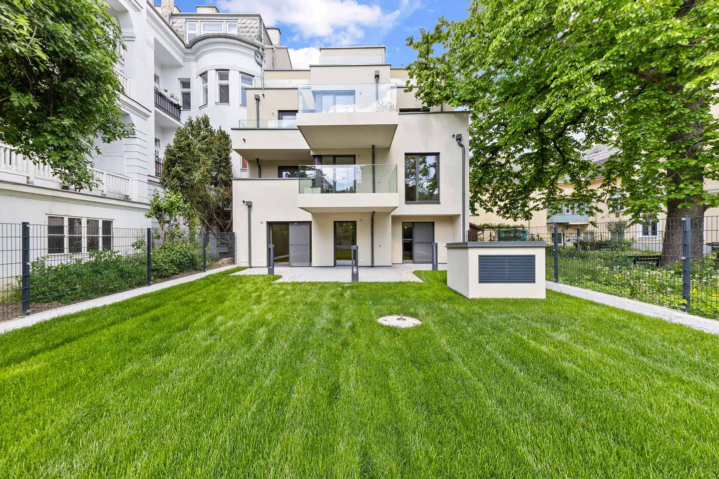 Modern, three-story cream building with glass balconies and a large green lawn, surrounded by a black metal fence.