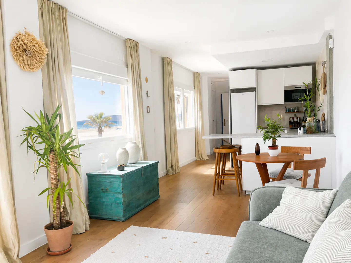 Bright, open-concept living space with a view of the beach. Features a teal chest, wood floors, and a modern white kitchen.