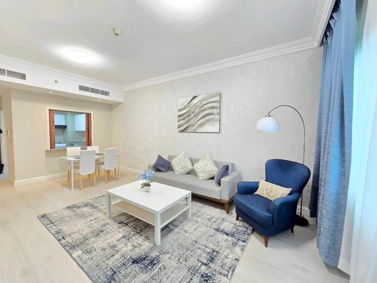 Living room with gray sofa, blue armchair, white table, and dining set. Blue patterned rug on light wood floor. Neutral walls with art.