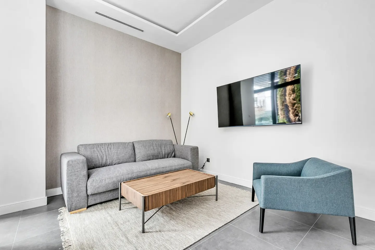 Modern living room with gray sofa, blue chair, and wood coffee table on a light rug. A TV hangs on the white wall.