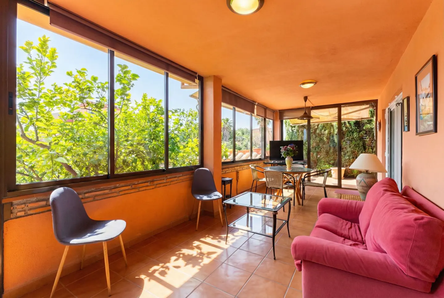 Bright sunroom with orange walls, tile floor, and large windows showing green trees. A pink sofa, chairs, and a glass table furnish the space.