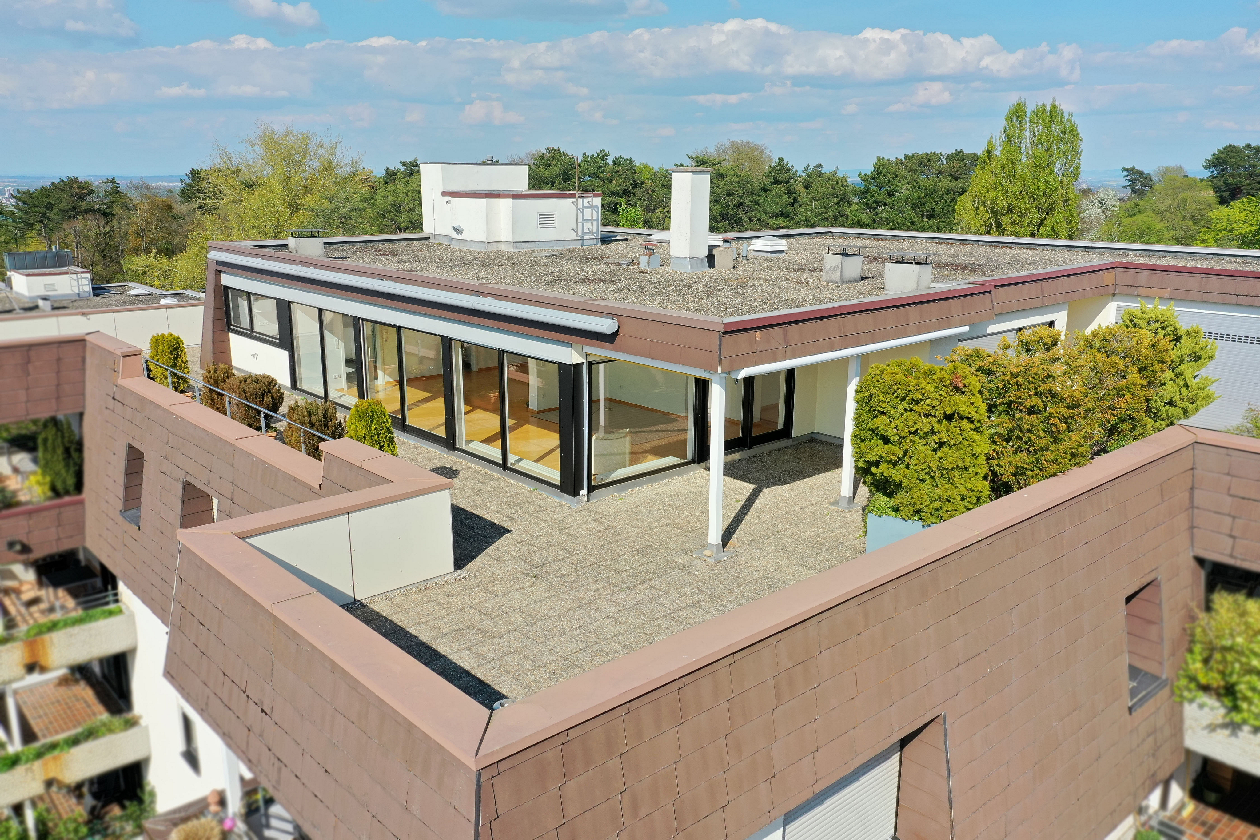 Aerial view of a modern building with a rooftop terrace, brown trim, and floor-to-ceiling windows. Trees and blue sky in the background.