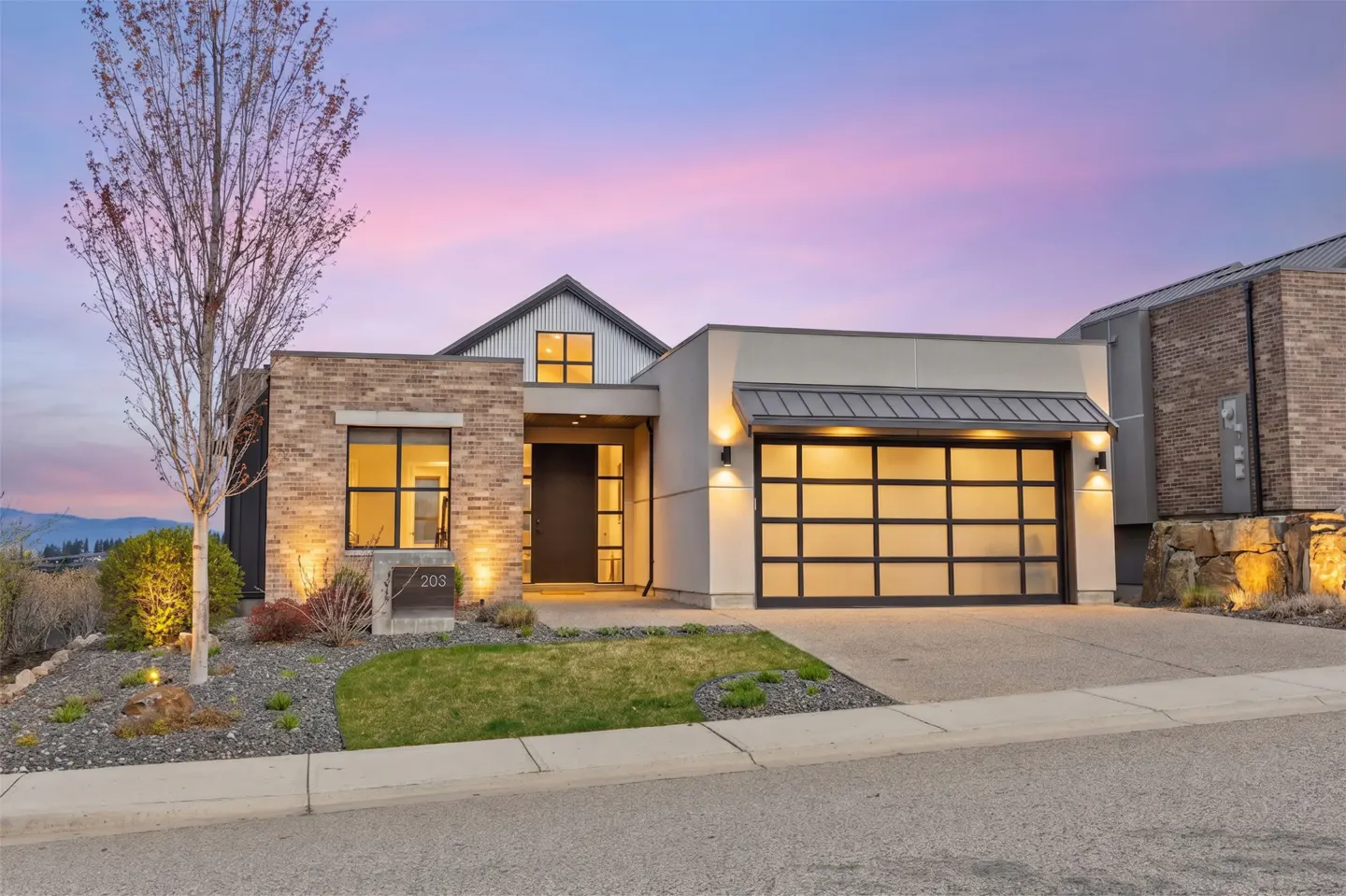 Modern home exterior at dusk. Brick and stucco facade, glass garage door, and manicured lawn. Pink and purple sky.