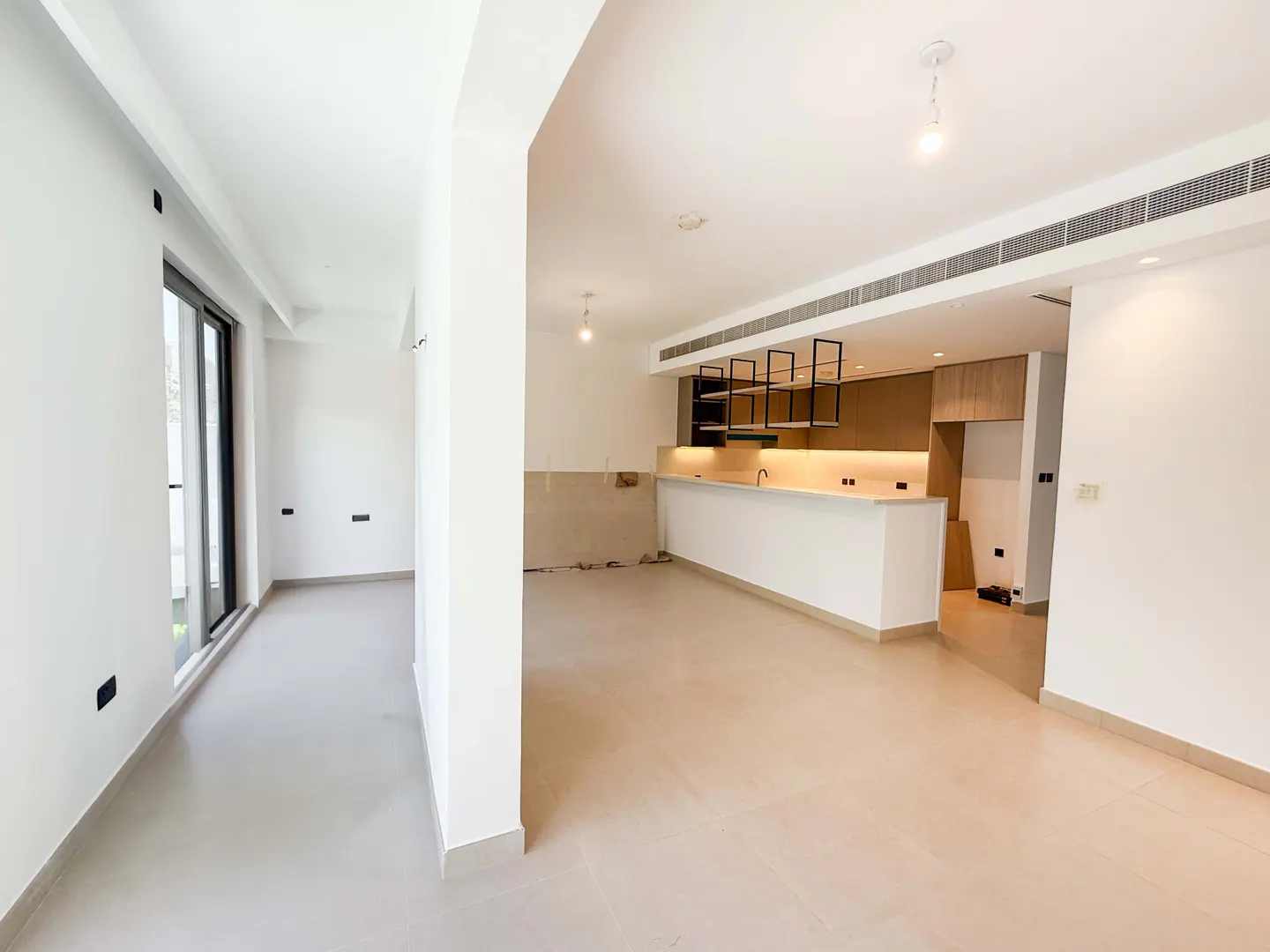 Bright, empty room with white walls and light beige floors. A kitchen area with a white island and wood cabinets is visible in the background. A large window lets in natural light.