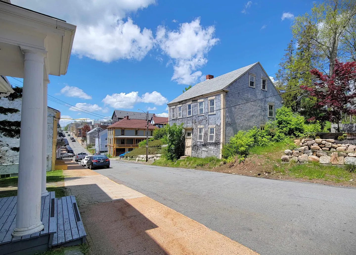 Street view of a gray clapboard house on a hill, with cars parked along the street under a blue sky with white clouds.