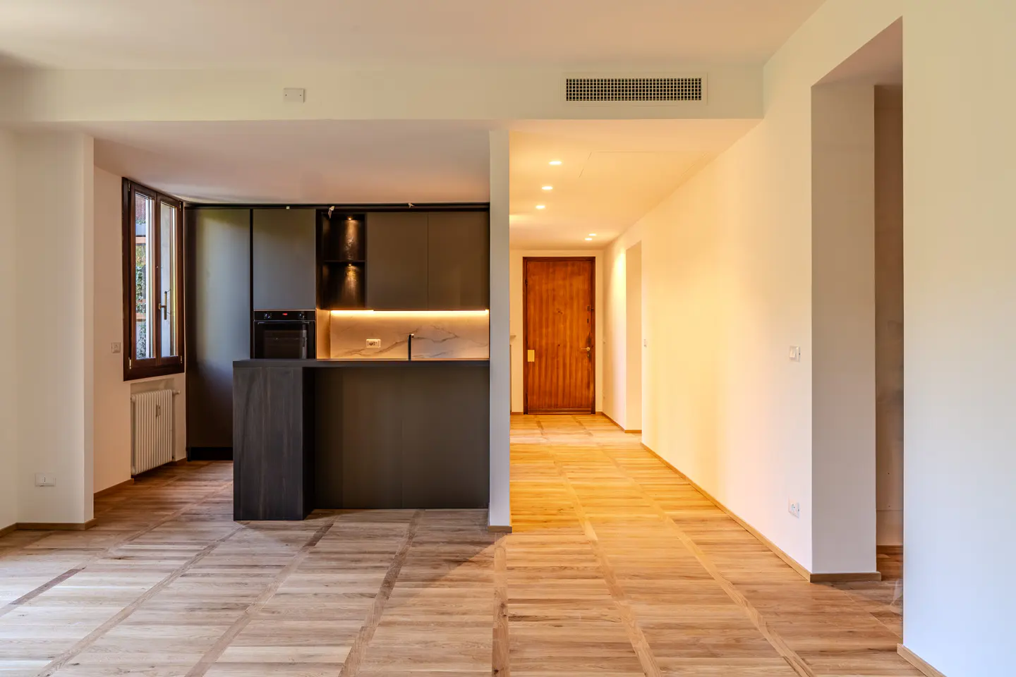 An interior view of a modern apartment with a kitchen, hallway, and wood floors. The kitchen has dark cabinets and a marble countertop. The hallway has a wooden door.