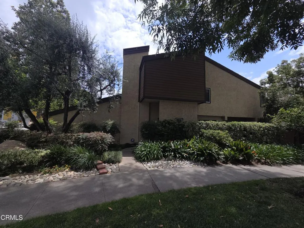 A two-story beige house with brown trim and a chimney, surrounded by lush green landscaping.