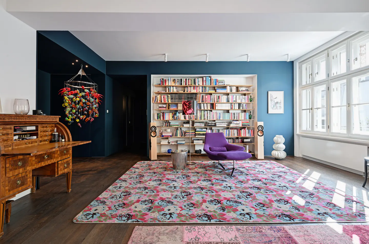 A living room with dark blue walls, a large bookshelf, a purple chair, and a floral rug. A wooden desk is on the left.