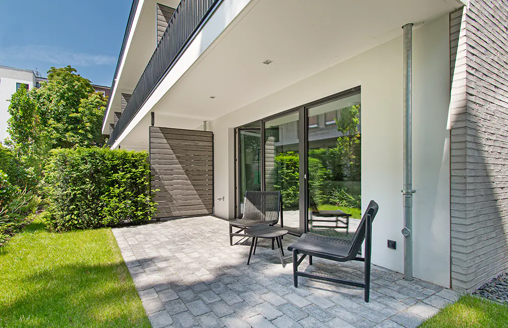 Modern patio with gray brick flooring, two black chairs, and a small table. Sliding glass doors reflect green foliage. A green lawn is visible.