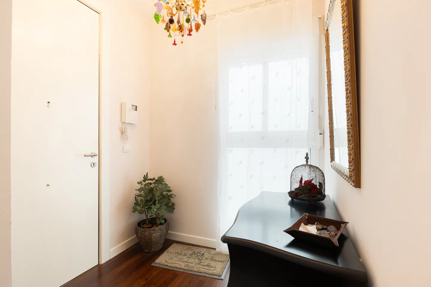 Entryway with a white door, dark wood floors, and a black console table. A colorful chandelier hangs above, and a gold-framed mirror is on the wall.
