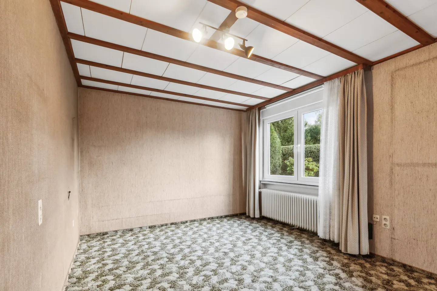 Empty room with beige textured walls, patterned carpet, and a window with white curtains. The ceiling has wooden beams and a track lighting fixture.