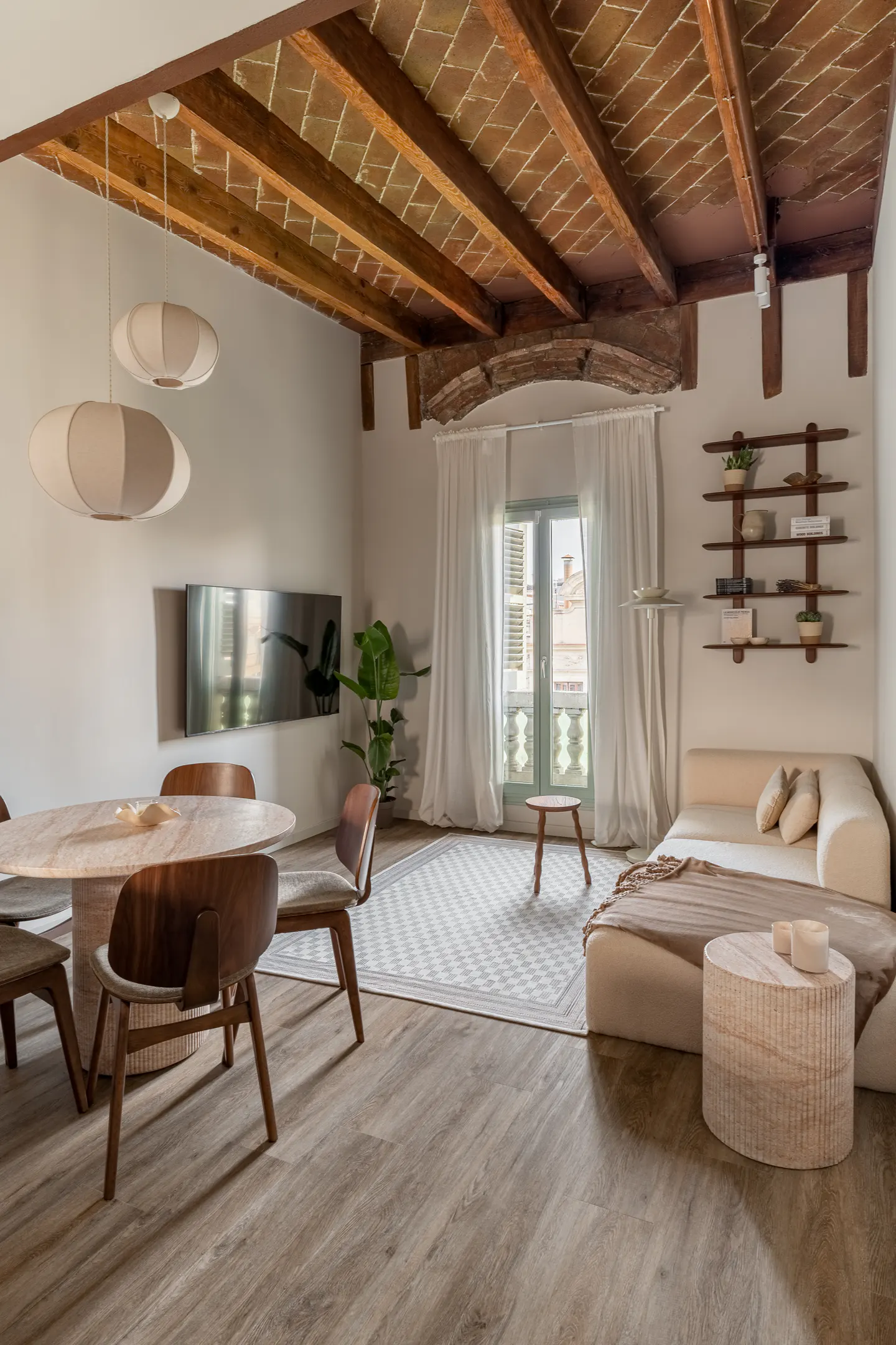 Bright living room with wood floors, exposed brick ceiling, and white walls. A round table and chairs sit near a TV, while a sofa and shelves are by a window.