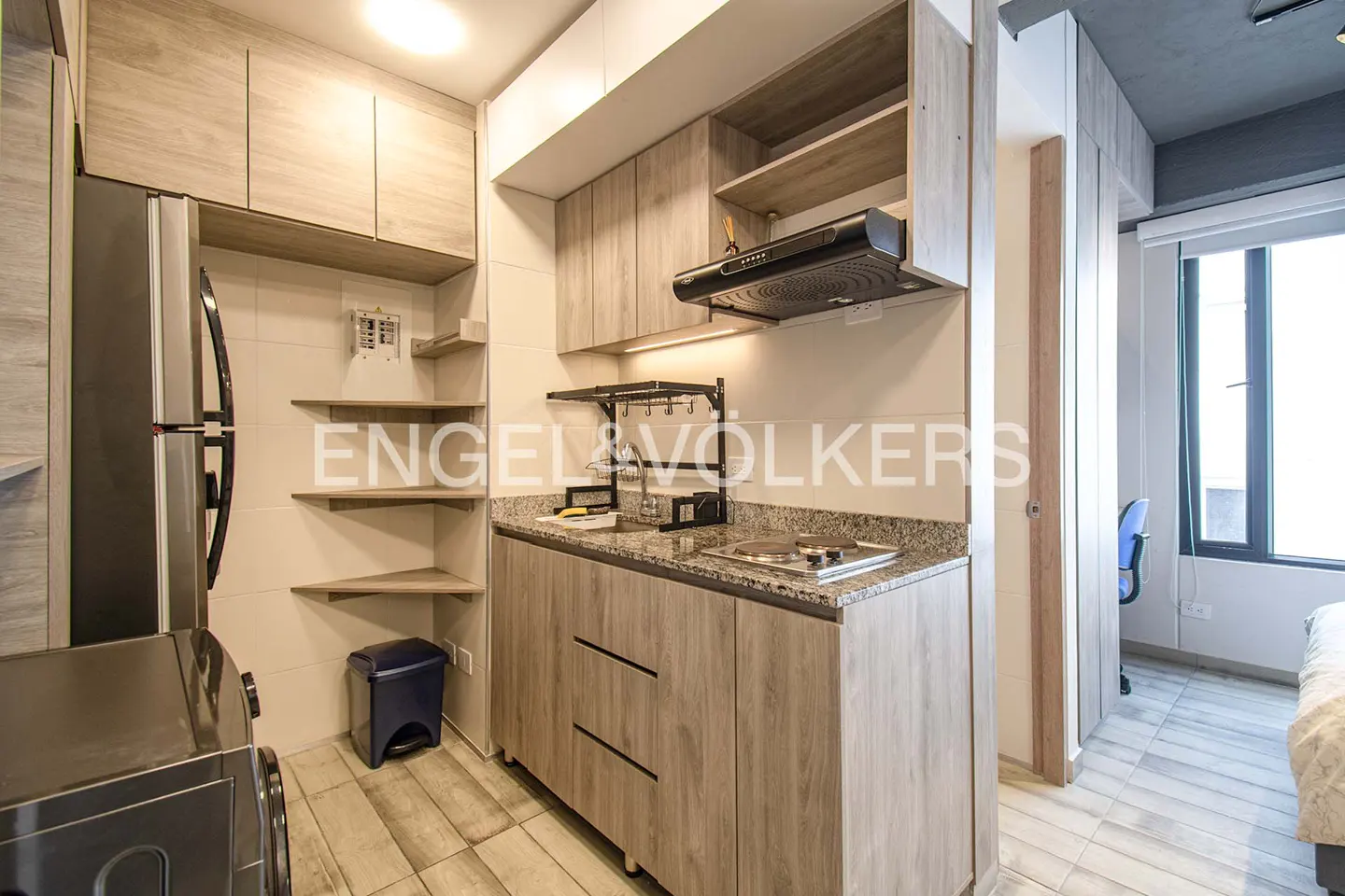 A compact kitchen with light wood cabinets, granite countertops, and stainless steel appliances. A blue trash can sits near open shelving.