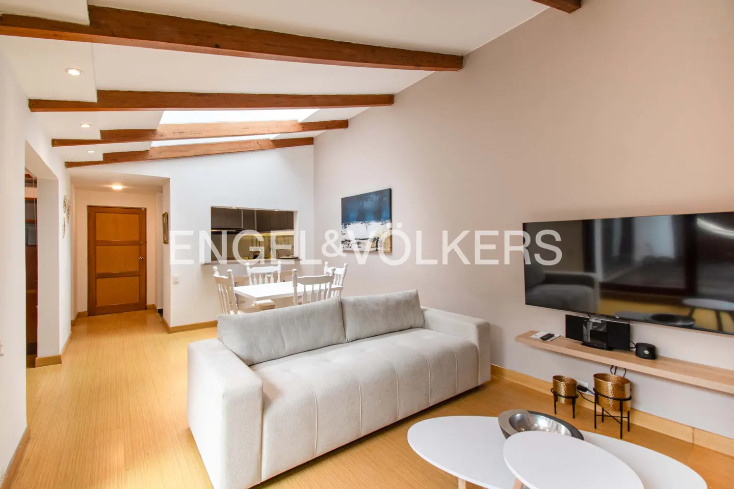 Living room with white sofa, TV, and wooden beams on the ceiling. A dining table is visible in the background.