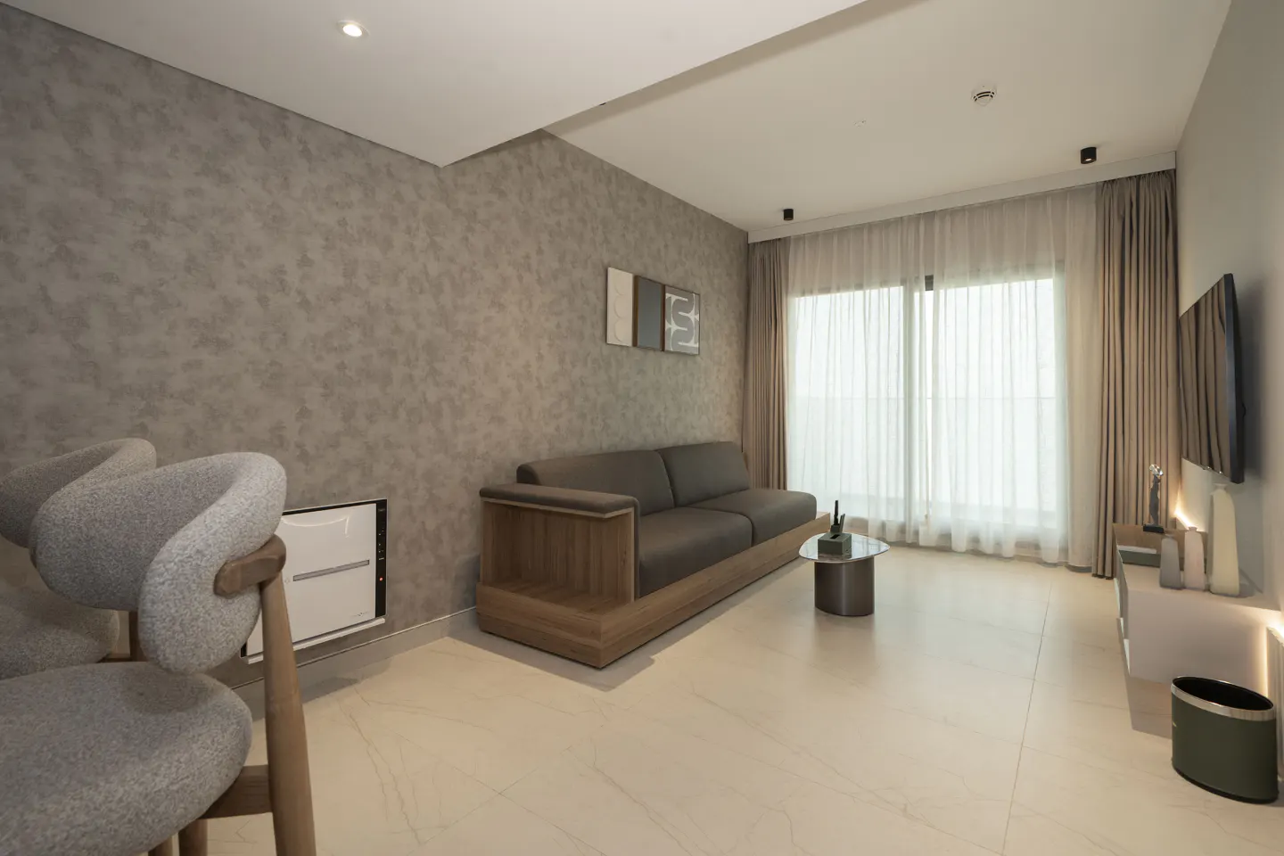 Modern living room with gray sofa, round glass table, TV, and gray chairs on a light marble floor. Neutral tones and natural light.