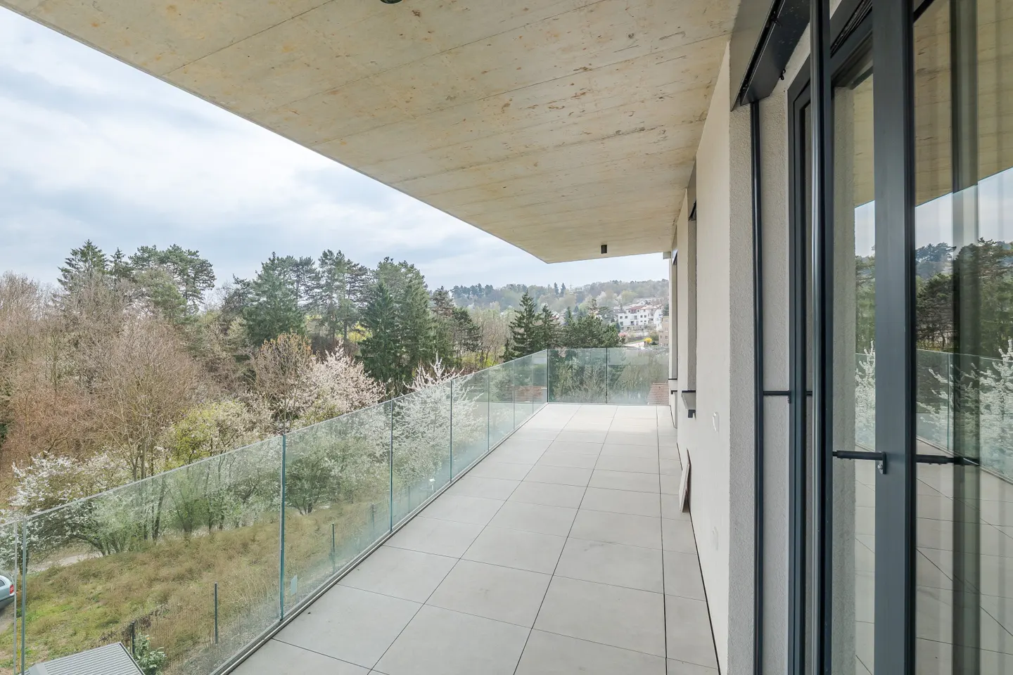 A modern balcony with glass railings overlooks a lush green landscape with trees and blooming white flowers. Gray tiled floor and sliding glass doors.