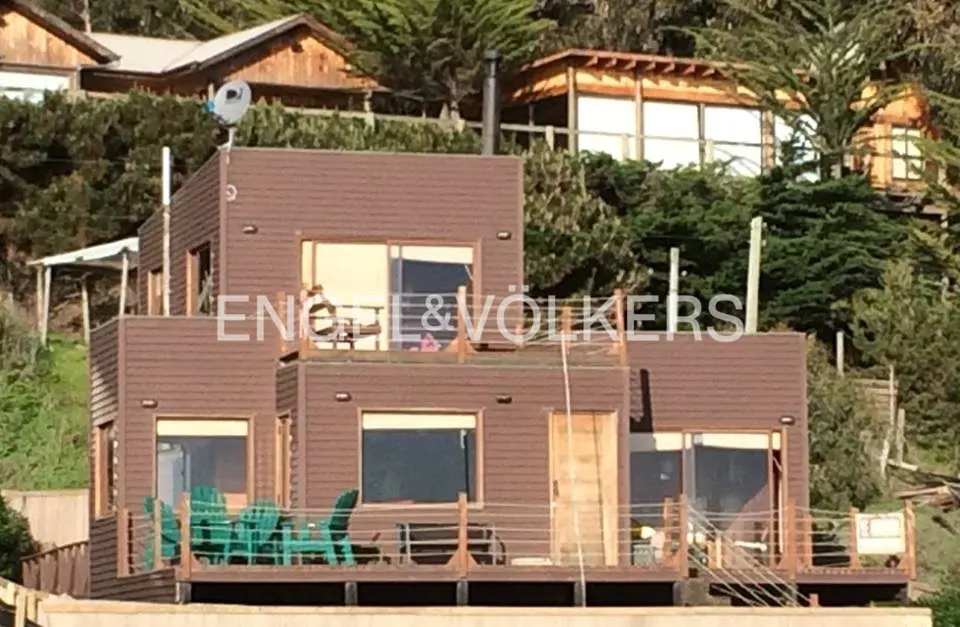 Modern two-story brown wooden house with a deck, large windows, and green chairs, set against a hillside with trees.