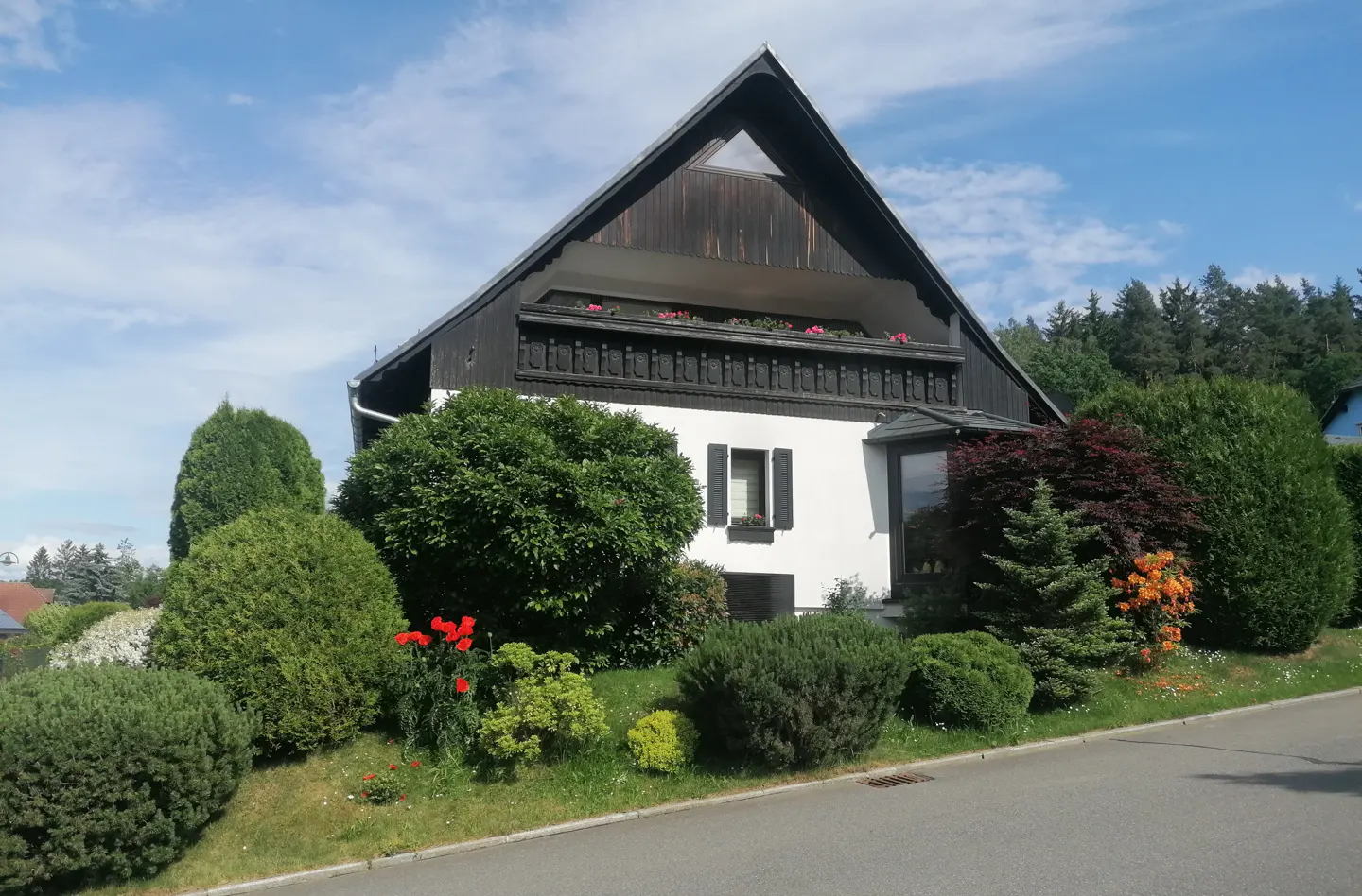 A white house with a black roof and trim is surrounded by green bushes and trees. Red flowers grow near the house.
