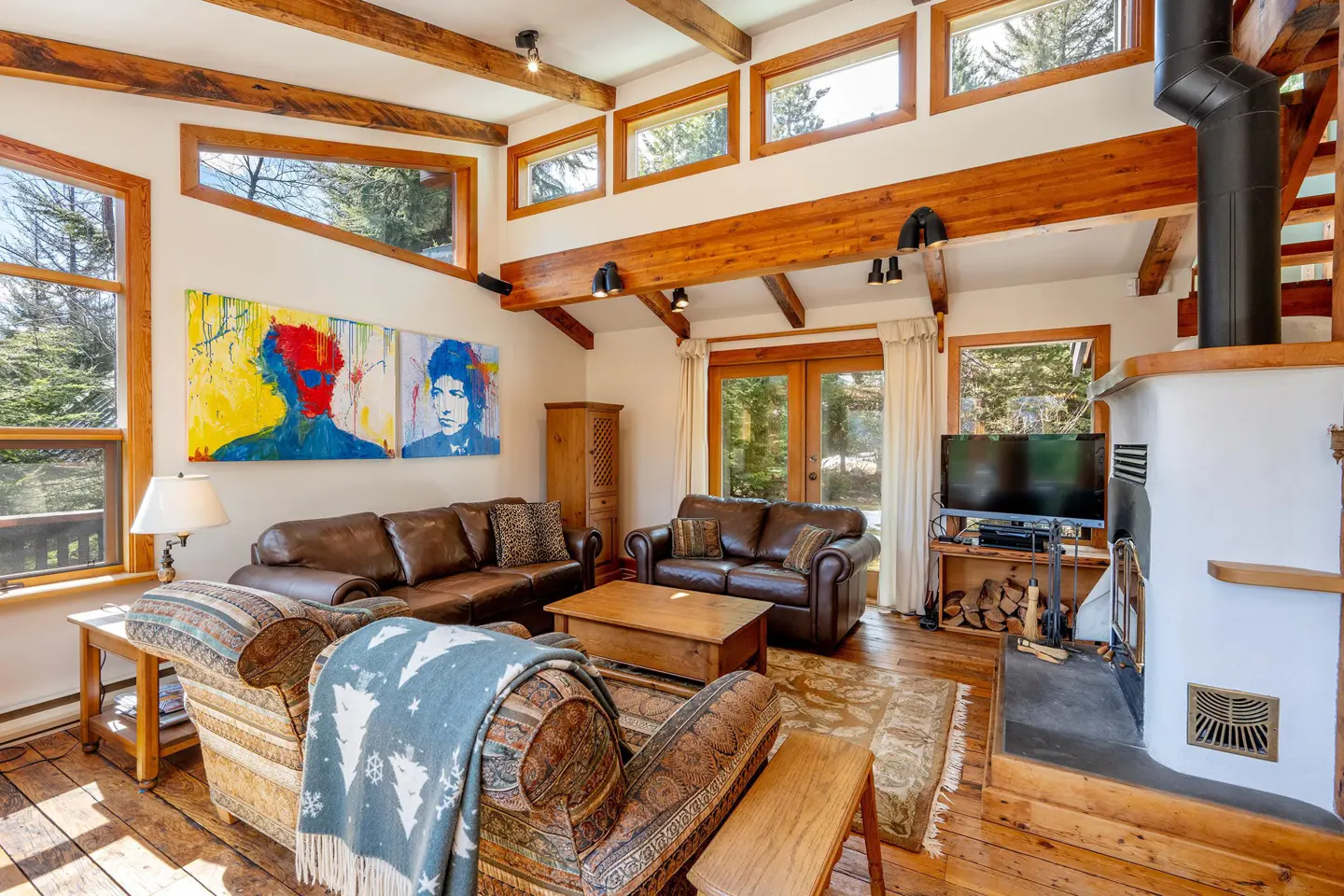 Living room with wood floors, exposed beams, and a fireplace. Two brown leather sofas face a patterned armchair with a blue blanket. Artwork on the wall.