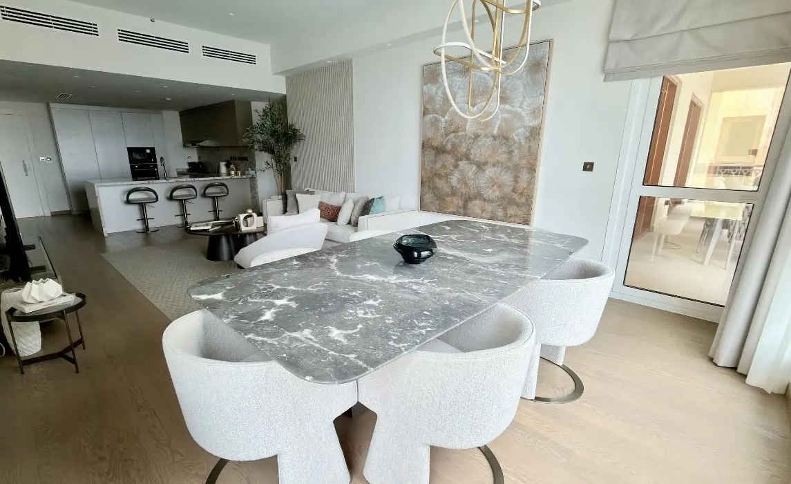 Modern dining area with a grey marble table, white curved chairs, and an open-plan kitchen in the background.