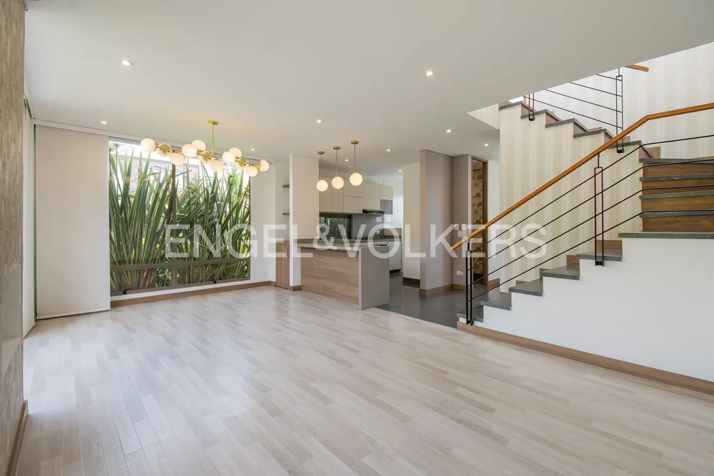 Modern open-plan living area with light wood floors, a kitchen island, globe pendant lights, and a wooden staircase.