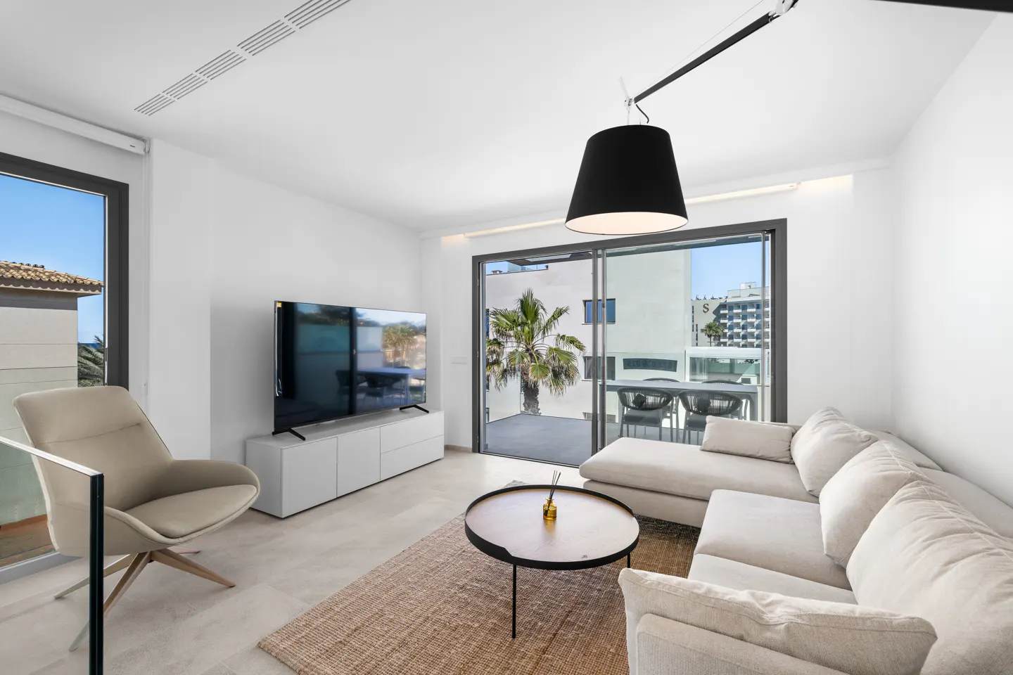 Bright living room with white walls, a beige sectional sofa, a round coffee table, and a TV. A palm tree is visible through the sliding glass doors.