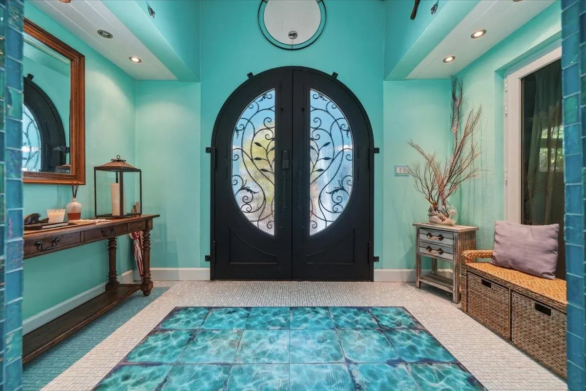 Entryway with teal walls, a black arched door with decorative ironwork, and a blue tile rug. A wooden table and bench sit on either side.