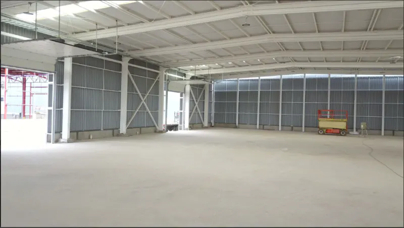 An empty warehouse with a concrete floor, metal walls, and a white ceiling. An orange scissor lift is parked near the back wall.